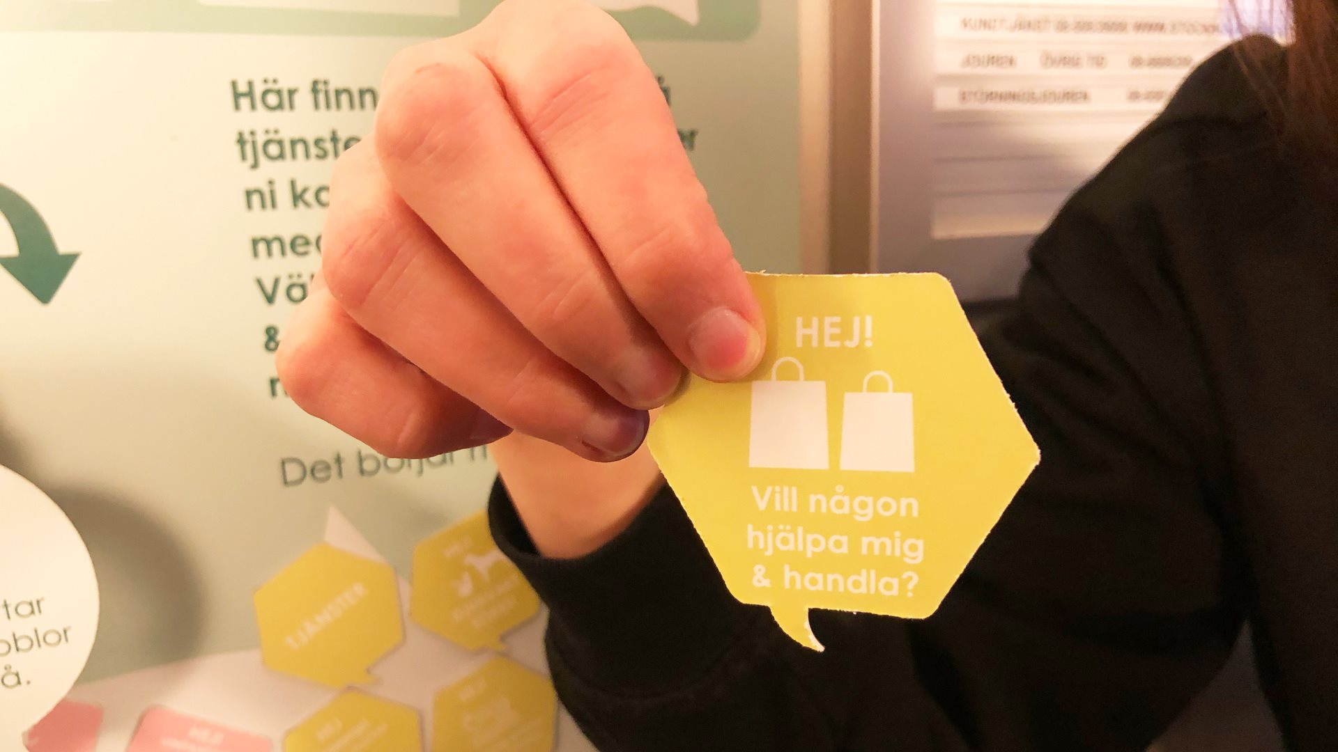 Hand håller liten bricka med texten "Hej! Vill någon hjälpa mig handla?"